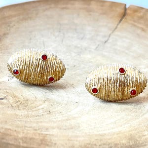 May include: A pair of oval-shaped gold-toned earrings with a textured, ribbed surface. Each earring is accented with three small, round, deep red stones embedded into the metal, creating a distinct contrast against the metallic gold finish.