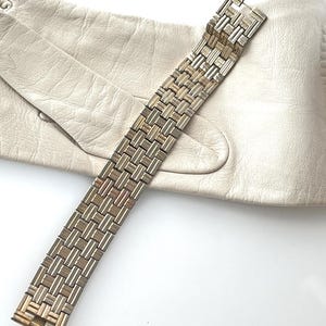 May include: A silver-toned bracelet with a woven design, set against a cream leather glove. The bracelet is composed of rectangular links, giving a textured look. The glove provides a soft, neutral backdrop, accentuating the bracelet's metallic sheen.