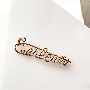 May include: A gold-colored wire name pin that reads "Carlean" in cursive script. The pin is made of a single piece of wire, with the letters connected. The pin is on a white surface.