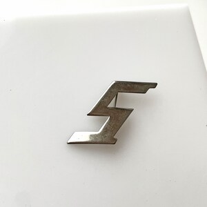 Puede incluir: Un broche de metal plateado con forma de rayo estilizado. El broche tiene un diseño geométrico con ángulos agudos y un pequeño elemento que sobresale en la parte superior. El broche está sobre un fondo blanco.