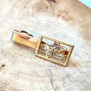 May include: A gold-toned tie clip featuring a detailed relief of a vintage car within a rectangular frame. The clip has a classic design, with a secure clasp for attaching to a tie. The car's wheels and body are clearly defined.