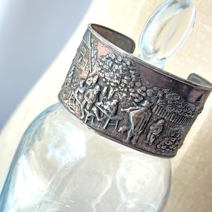 May include: A wide silver cuff bracelet featuring a detailed embossed relief scene of figures gathered outdoors near a house and trees. The metal has a darkened patina in the recessed areas, highlighting the intricate landscape and human figures.