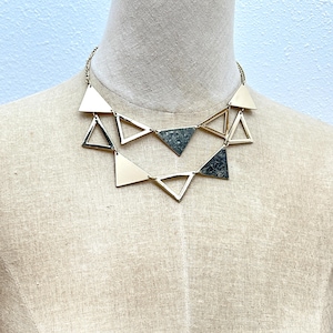 Op de afbeelding: Een goudkleurige metalen ketting met twee lagen geometrische driehoekige hangers. Het ontwerp bevat een mix van effen goudkleurige driehoeken en getextureerde, donkergrijze gespikkelde driehoeken verbonden door dunne goudkleurige metalen kettingen.