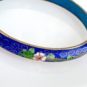 May include: A blue cloisonné bangle bracelet with floral and cloud designs. The bracelet features a gold-colored metal band and is decorated with pink, green, and white flowers. The bracelet has a diameter of approximately 2.5 inches.