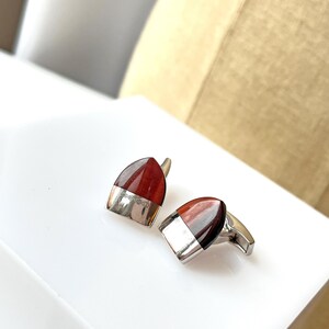 May include: A pair of cufflinks with a silver base and a reddish-brown, rounded top. The top portion of each cufflink is a polished, reddish-brown color. The bottom half is silver. The cufflinks are on a white surface.