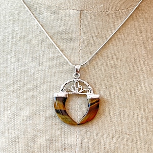 May include: A silver-toned necklace featuring a pendant with two curved, polished tiger's eye stones in shades of brown and gold. The stones are set in a silver-colored metal frame with an ornate, filigree-style arch at the top and a thin chain.