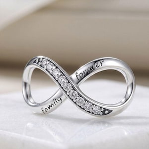 May include: A silver-toned infinity charm featuring a row of clear, round-cut stones along one side. The metal surface is engraved with the word family on the lower left curve and the word forever on the upper right curve of the infinity symbol.