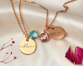 Peut inclure: Deux colliers en or avec des médaillons de nom personnalisés et de petits pendentifs en pierres précieuses. Les colliers sont sur un fond en tissu blanc. Les médaillons portent les noms "Chloe" et "Jasmin".