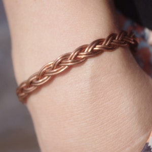 May include: A copper-colored braided bracelet. The bracelet is made of multiple strands woven together, creating a textured, rope-like appearance. The jewelry is worn on the wrist.