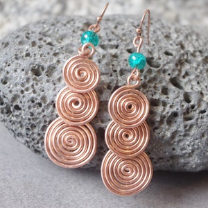 Pode incluir: Um par de brincos de ouro rosa com três círculos em espiral e uma conta verde-azulada na parte superior de cada brinco.