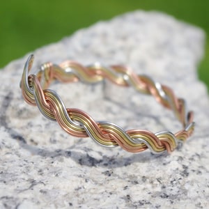 Könnte beinhalten: Ein geflochtenes Armreif-Armband aus drei verschiedenen farbigen Drähten: Silber, Gold und Kupfer. Das Armband liegt auf einem grauen Stein.