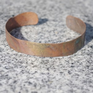 May include: A copper cuff bracelet with a weathered, aged appearance. The open-ended bracelet has a slightly curved shape and shows variations in color, including shades of brown, gold, and hints of green. The bracelet is approximately 2 inches wide.