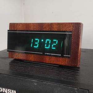 May include: A vintage digital clock with a wooden frame displays the time 13:02 in bright green digits. The clock face is black with a silver border, and the wooden frame has a warm, reddish-brown finish. The word "электроника" is printed on the clock face.
