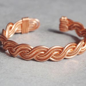 Könnte beinhalten: Ein Armband aus Kupferdraht mit geflochtenem Design. Das Armband besteht aus mehreren Drähten, die miteinander verflochten sind, um ein einzigartiges und kompliziertes Muster zu schaffen.