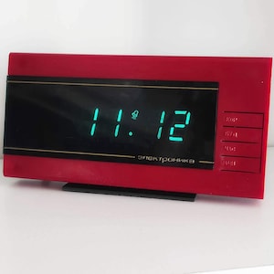 May include: A red digital clock with a green display showing the time 11:12. The clock has a black face and a silver trim. The clock is labelled "Электроника" in Cyrillic.