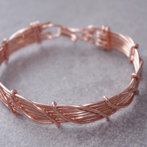 Puede incluir: Un brazalete de alambre envuelto en oro rosa. El brazalete está hecho de múltiples hilos de alambre que se tejen juntos para crear un diseño único e intrincado.