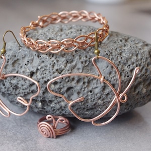 Könnte beinhalten: Ein Set aus Kupferdraht-Schmuck mit einem geflochtenen Armband, einem Paar Schmetterlings-Ohrringen und einem Spiralring. Der Schmuck wird auf einem grauen Stein präsentiert.