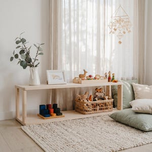 Peut inclure: Une étagère en bois clair à deux niveaux présente des jouets en bois, une impression encadrée d'arc-en-ciel et un vase blanc avec de la verdure. Un panier tressé contient d'autres jouets et des coussins reposent sur le sol. Un mobile géométrique est suspendu au plafond.