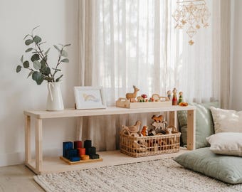 May include: A light wood, two-tiered shelf unit displays wooden toys, a framed rainbow print, and a white vase with greenery. A woven basket holds more toys, and pillows rest on the floor. A geometric mobile hangs from the ceiling.