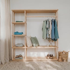 Pode incluir: Um guarda-roupa de madeira clara com várias prateleiras e uma barra para pendurar roupas. O guarda-roupa contém roupas dobradas, travesseiros, sapatos e roupas penduradas em tons de azul, verde e bege. Uma cesta de vime com brinquedos de madeira está no chão.