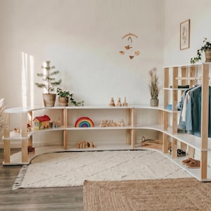 May include: A white and wood-toned shelving unit with a curved design, filled with wooden toys, plants, and hanging clothes. The room has a neutral color palette with a rug and a mobile hanging from the ceiling.