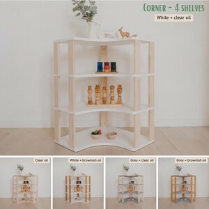 May include: A white and natural wood corner shelf unit with four shelves. The text "CORNER - 4 SHELVES" and "White + clear oil" are visible. The shelves are holding wooden toys and decorative items.