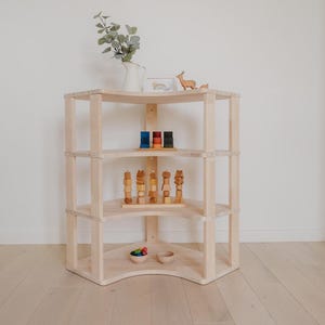 May include: A three-tiered, light-colored wooden corner shelf unit. The shelves display wooden toys, a vase with greenery, and small decorative items. The unit is placed against a white wall and light wood floor.