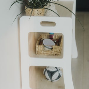 The Montessori shelf is photographed from the side to show the side of the shelf. It is cut to the shape of the shelves, lightweight, minimalist, with handles cut into the top to make it easy to move the Montessori shelf.