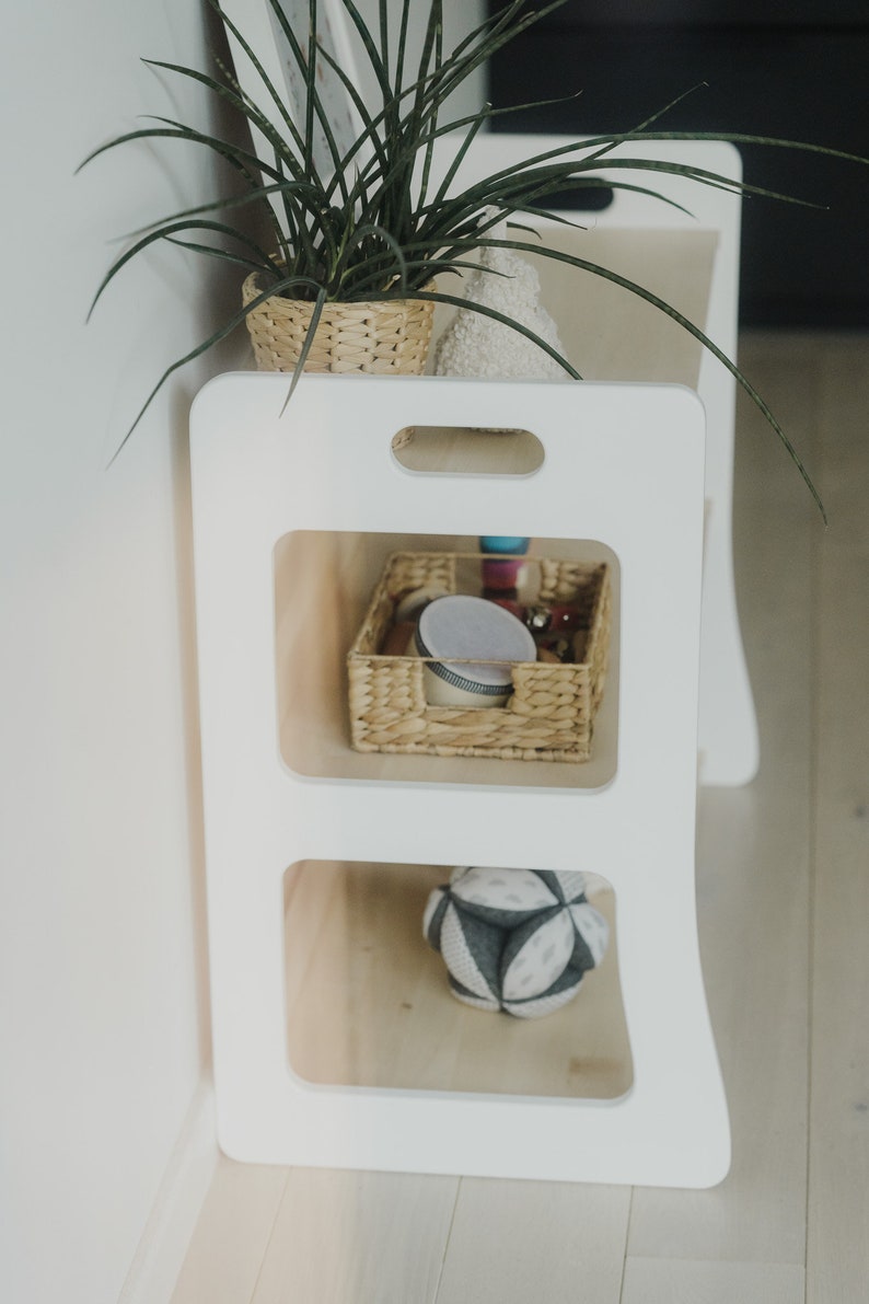 The Montessori shelf is photographed from the side to show the side of the shelf. It is cut to the shape of the shelves, lightweight, minimalist, with handles cut into the top to make it easy to move the Montessori shelf.