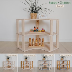 May include: A three-tiered corner shelf unit made of light-colored wood. The shelves are curved and hold wooden toys and a small plant in a woven basket. The text "CORNER - 3 SHELVES" and "Clear oil" are visible.