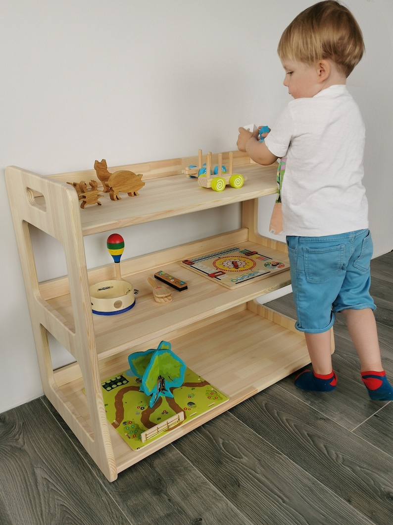 The child takes toys from the Montessori shelf. This Montessori shelf is made of natural wood, untreated. The natural wood is light and the structure of the wood is visible. Montessori shelf of 3 shelves, dimensions: Height 66, width 81, depth 42 cm.