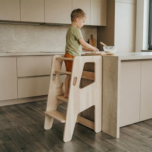 May include: A light-coloured wooden step stool with a safety rail and two steps. The step stool is designed to help a child reach a kitchen work surface. The child is wearing an olive green top and orange shorts.