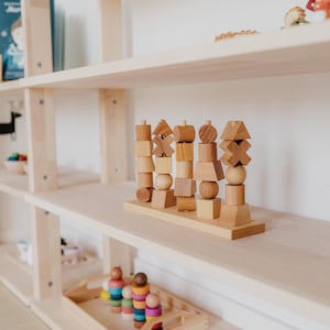 May include: Wooden educational toy with various geometric shapes stacked on a wooden base. The toy is made of light-colored wood and sits on a light-colored wooden shelf. Other wooden toys and books are visible on the shelves.