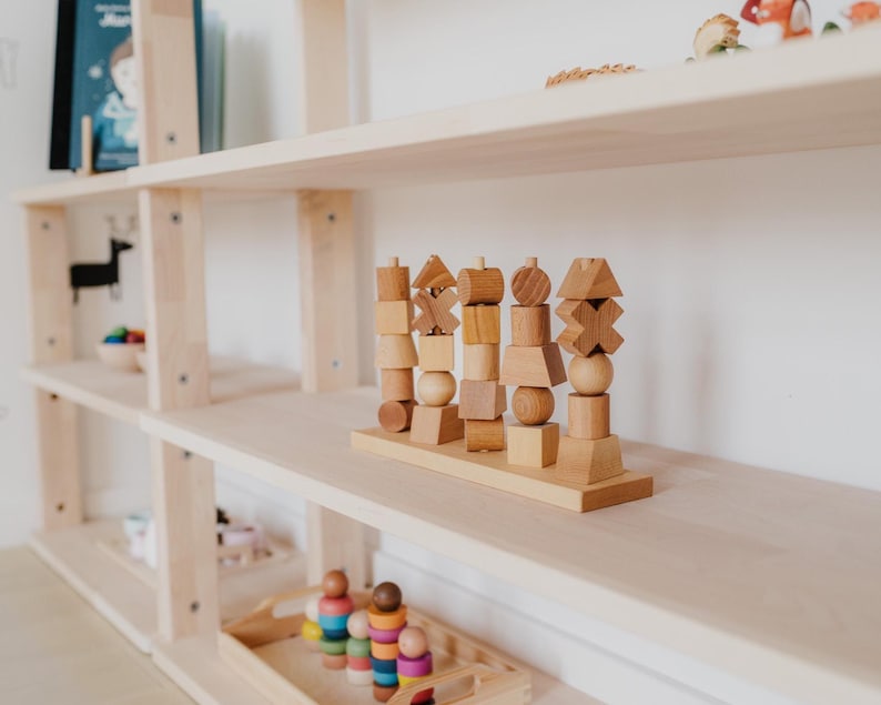 May include: Wooden educational toy with various geometric shapes stacked on a wooden base. The toy is made of light-colored wood and sits on a light-colored wooden shelf. Other wooden toys and books are visible on the shelves.