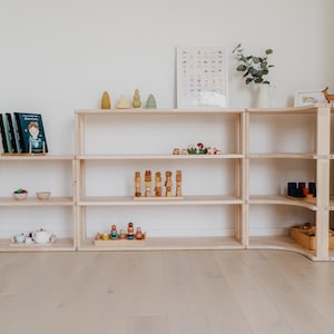 May include: A light-colored wooden shelving unit with multiple shelves, arranged in a corner configuration. The shelves are filled with various items, including books, wooden toys, and decorative objects. The unit is placed against a white wall and light wood floor.