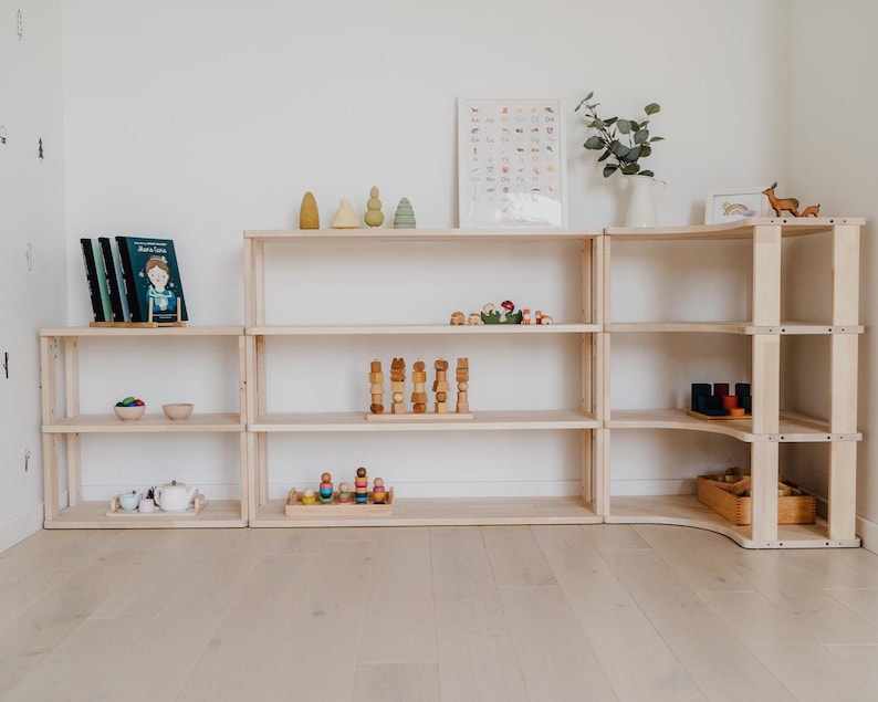 May include: A light-colored wooden shelving unit with multiple shelves, arranged in a corner configuration. The shelves are filled with various items, including books, wooden toys, and decorative objects. The unit is placed against a white wall and light wood floor.