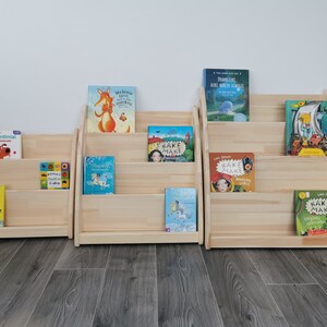 The photo shows 3 Montessori bookshelves of different sizes, all untreated. The bookcases are placed on the ground, sturdy and stable because they are wide and deep, All corners are rounded. Handles cut into the side panels make it easy to move them.