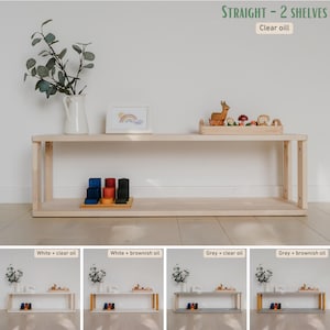 May include: A light-colored wooden shelf unit with two shelves. The unit is rectangular and has a natural wood finish. The top shelf holds a white vase with greenery, a framed picture, and a wooden tray with toys. The text "STRAIGHT - 2 SHELVES" and "Clear oil" are visible.