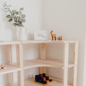 May include: A light-colored wooden shelf unit with a corner design. The shelves display a white pitcher with greenery, a framed rainbow print, and wooden deer figurines. A wooden block set is on the lower shelf.