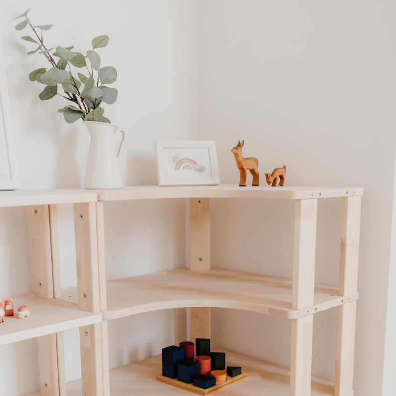 May include: A light-colored wooden shelf unit with a corner design. The shelves display a white pitcher with greenery, a framed rainbow print, and wooden deer figurines. A wooden block set is on the lower shelf.