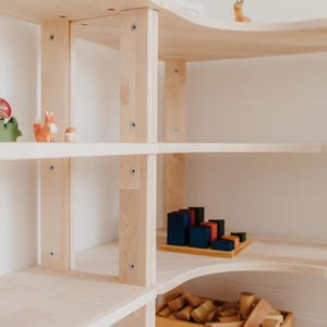 May include: A light-colored wooden bookshelf with multiple shelves. The shelves display wooden toys, including a fox figurine, a mushroom, and geometric blocks. The bookshelf has a curved top and is against a white wall.