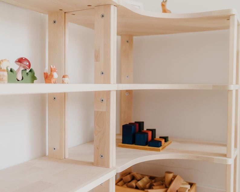 May include: A light-colored wooden bookshelf with multiple shelves. The shelves display wooden toys, including a fox figurine, a mushroom, and geometric blocks. The bookshelf has a curved top and is against a white wall.