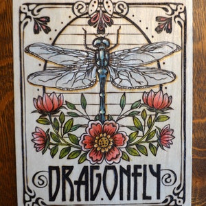 May include: A woodburned art piece with a dragonfly in the center, surrounded by flowers and leaves. The words "Dragonfly" are written below the dragonfly.