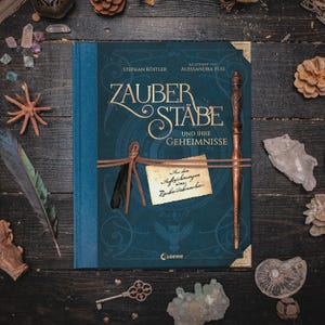 Könnte beinhalten: Ein Hardcover-Buch mit dem Titel "Zauberstäbe und ihre Geheimnisse" (Zauberstäbe und ihre Geheimnisse) in Blau und Gold, mit einem Zauberstab und einer kleinen Notiz, die daran befestigt ist. Das Buch ist von Kristallen, Federn und anderen dekorativen Gegenständen auf einer dunklen Holzoberfläche umgeben.