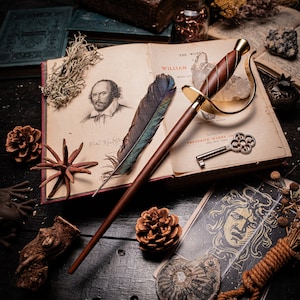 May include: A gold-toned decorative sword with a brown wooden handle and a feather quill. The sword is lying on top of an open book with the title "The Works of William Shakespeare" printed on the cover. The book is surrounded by other objects, including a small wooden box, a pine cone, and a feather.
