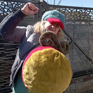 Op de afbeelding: Een vrouw met een groene en rode hoed houdt een groot, rond, pluche speelgoed vast. Het speelgoed is bruin en geel met een rode accent.