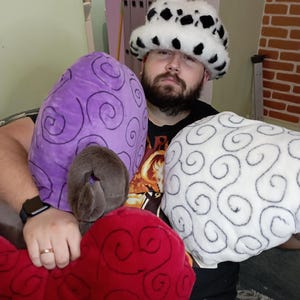 Könnte beinhalten: Eine Person trägt einen Hut mit weißem und schwarzem Muster und hält drei Plüschtiere. Die Spielzeuge sind gehirnförmig und haben ein Wirbelmuster. Die Spielzeuge sind lila, weiß und rot.