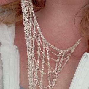May include: A multi-strand silver-colored necklace featuring a web-like design composed of numerous delicate, interconnected fine metal chains that drape across the chest area, creating a complex, layered, and open-work geometric pattern.