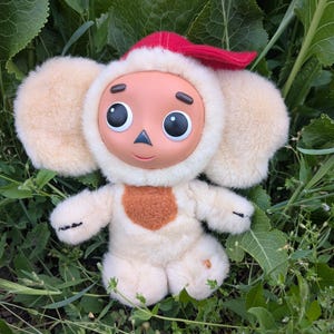 Op de afbeelding: Een pluchen speelgoedfiguur met een crèmekleurig lichaam, grote pluizige oren en een rode hoed. Het speelgoed heeft een rond gezicht met grote zwarte ogen, een kleine neus en een lachende mond.