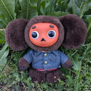 May include: A plush toy character with a brown furry head, large ears, and a round orange face with big eyes. It wears a denim jacket and sits in green grass. The toy has brown fur on its arms and legs.